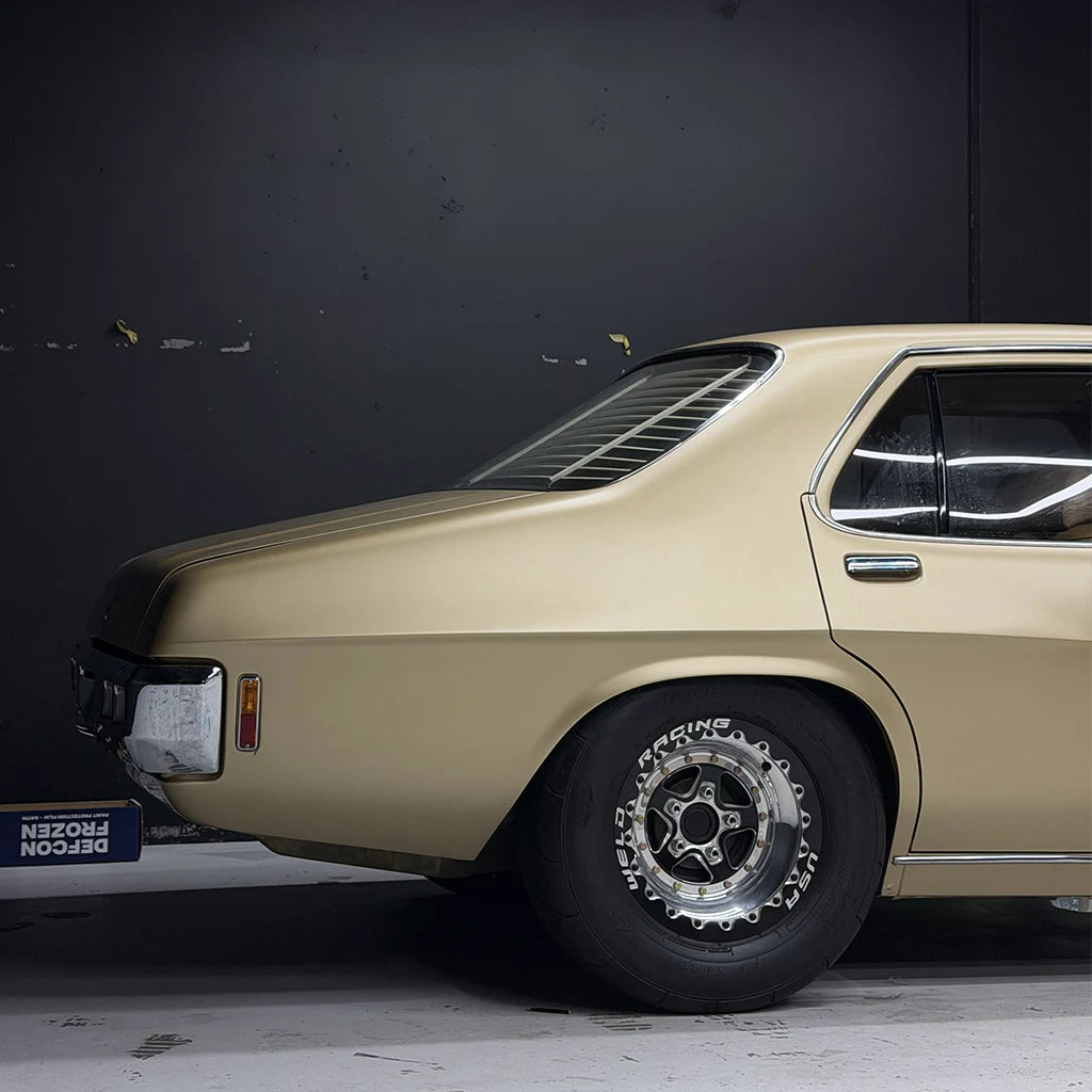 A side view of a classic car in a matte champagne gold finish, featuring a black vinyl roof, a vented rear window, and custom multi-spoke wheels with 'Racing' lettering, set against a dark gray studio background.