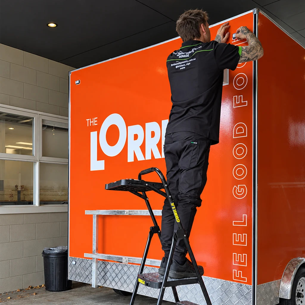 A man in black work clothes stands on a step ladder, applying a protective film or tape to the top corner of a large orange trailer or truck side panel. The panel has white text reading 'THE LORRI' and 'FEEL GOOD FOOD' in part of a larger slogan.